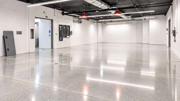 Large open multipurpose room with a light cream polished concrete floor, exposed ceiling services, and red pendant fixtures overhead